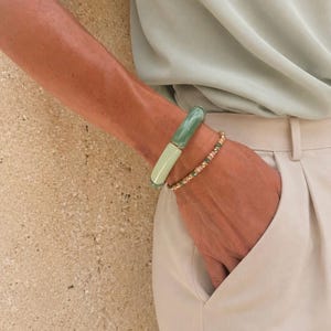 May include: A two-toned bracelet featuring a smooth, cylindrical dark green segment and a lighter mint green segment, worn alongside a delicate beaded bracelet with small metallic accents, paired with a light beige shirt and trousers.