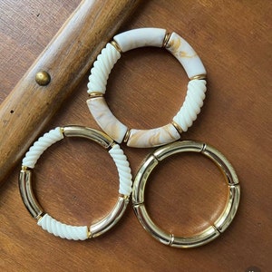 Könnte beinhalten: Drei Armbänder, eines mit einem weißen und braunen Marmormuster und zwei mit einem goldenen Metall- und weißen gedrehten Design.