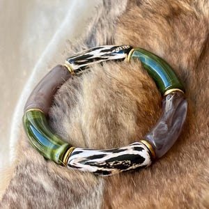 May include: A chunky bracelet featuring a mix of colorful beads. The bracelet includes green, brown, and white beads with gold-colored accents. The white beads have a black pattern. The bracelet is displayed on a textured, brown surface.