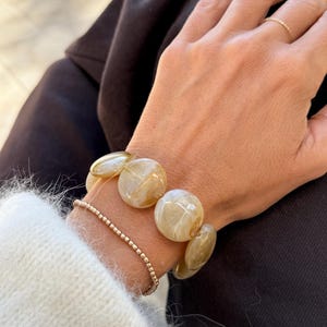 May include: A wrist adorned with two bracelets. One is a beaded gold bracelet, and the other is a chunky bracelet with large, translucent, light brown beads. The person is wearing a white sweater and black pants.