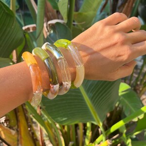 Pu&ograve; includere: Quattro braccialetti in resina colorati impilati su un polso. I braccialetti sono in tonalit&agrave; di verde, marrone, arancione e bianco.