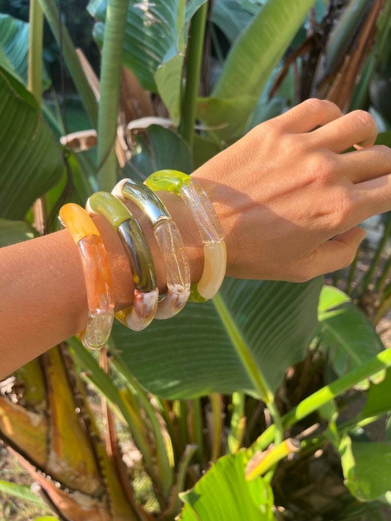 Pu&ograve; includere: Quattro braccialetti in resina colorati impilati su un polso. I braccialetti sono in tonalit&agrave; di verde, marrone, arancione e bianco.