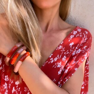 Könnte beinhalten: Ein rotes Kleid mit weißem Blumenmuster. Die Person trägt mehrere klobige, farbenfrohe Armbänder in Rot-, Rosa- und Beigetönen. Goldene Creolen sind ebenfalls zu sehen.