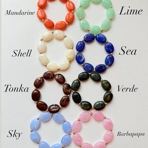 May include: An assortment of colorful beaded rings in various colors, including orange, green, white, blue, brown, and pink. Each ring is made of oval-shaped beads and features gold-colored accents. The rings are labeled with color names such as "Mandarine" and "Lime".