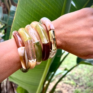 Pu&ograve; includere: Un set di cinque braccialetti colorati, impilati su un polso. I braccialetti sono in plastica trasparente e hanno un effetto marmorizzato. I colori sono marrone, verde, beige e bianco.