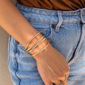 May include: Gold-toned bracelets and rings adorn a person's wrist and fingers. The bracelets include a plain band, a textured band, and a band with a clasp. The person is wearing blue jeans and a brown top.
