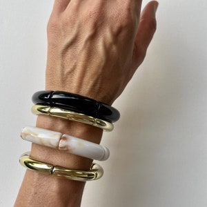 Könnte beinhalten: Vier klobige Armbänder in verschiedenen Farben und Materialien. Das oberste Armband ist schwarz, gefolgt von einem goldfarbenen Armband, einem weiß-cremefarbenen Marmorarmband und einem weiteren goldfarbenen Armband. Die Armbänder werden an einem Handgelenk getragen.