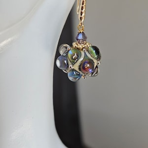 Könnte beinhalten: Ein goldener Ohrring mit einem Cluster aus irisierenden Glasperlen und einem violetten Kristall-Anhänger.