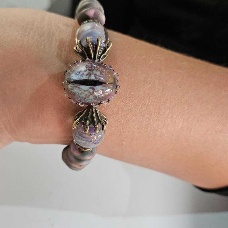 May include: A beaded bracelet featuring a central eye-shaped bead with a black pupil, surrounded by smaller, iridescent beads in shades of purple and gray. The bracelet has bronze-colored decorative elements and is designed to be worn on the wrist.
