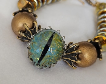 May include: A gold and green bracelet with a large, eye-shaped bead. The bead is made of glass and has a swirling pattern of green, blue, and gold. The bracelet is made of metal and has a decorative design.