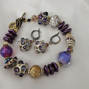 Puede incluir: Un brazalete y pendientes a juego de cuentas de vidrio morado y blanco. El brazalete presenta una variedad de cuentas de vidrio, incluyendo morado, blanco y dorado. Los pendientes son simples aros de plata con una sola cuenta de vidrio blanco.
