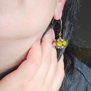 Peut inclure: Boucles d'oreilles créoles dorées avec une perle de verre jaune et noire suspendue. La perle est une sphère avec une surface texturée et de nombreux petits cercles jaunes et noirs.