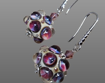 May include: A pair of silver dangle earrings with a white and purple glass bead. The beads are round and have a textured surface. The earrings are a unique and stylish accessory.