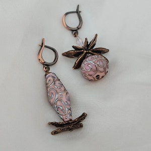 May include: A pair of mismatched dangle earrings featuring antique copper-toned metal findings and pale pink beads with silver-coloured swirling filigree patterns. One earring has a round bead with a star-shaped topper, while the other has a long bead.