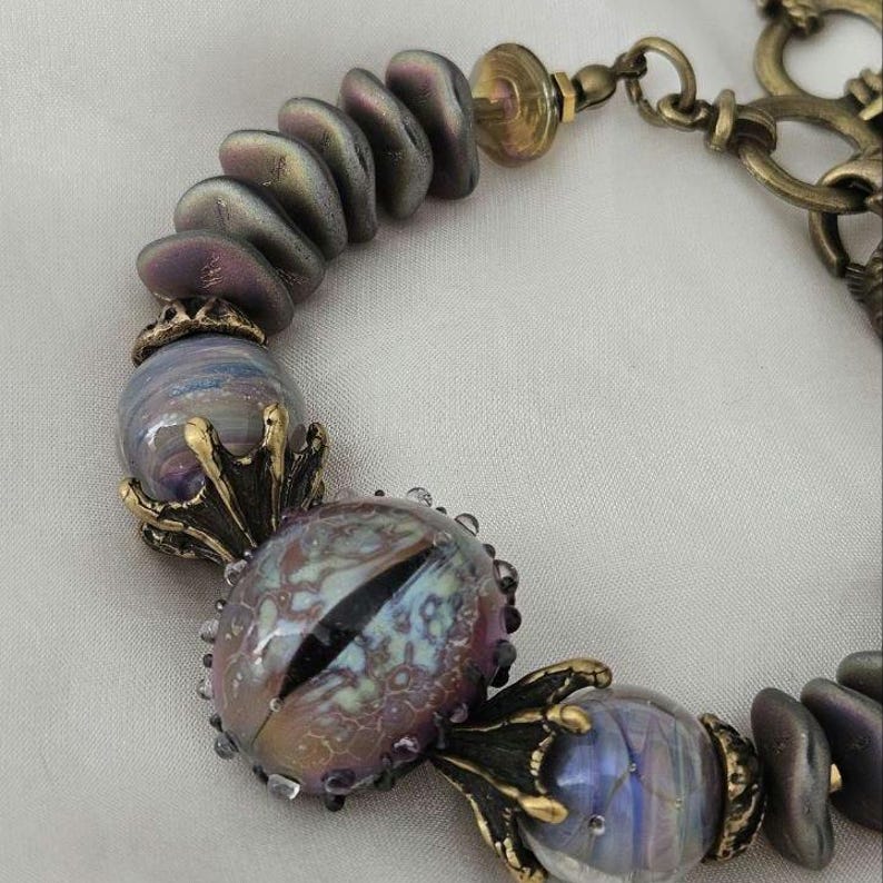 May include: A handcrafted bracelet featuring iridescent purple and blue glass beads. The focal point is a large, eye-shaped bead with a black slit pupil. The bracelet includes bronze-colored metal accents and a decorative clasp.