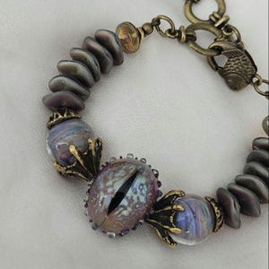 May include: A unique bracelet featuring iridescent, oval-shaped beads and a central eye-shaped bead with a black slit pupil. The bracelet includes gold-toned metal accents and a fish-shaped clasp, creating a fantasy-themed accessory.