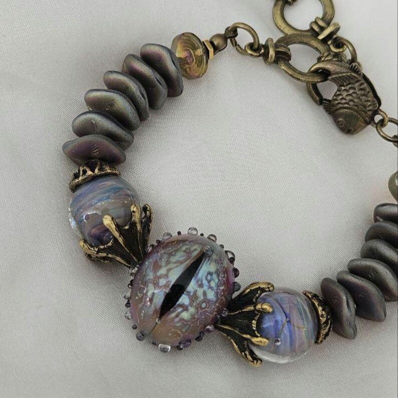 May include: A unique bracelet featuring iridescent, oval-shaped beads and a central eye-shaped bead with a black slit pupil. The bracelet includes gold-toned metal accents and a fish-shaped clasp, creating a fantasy-themed accessory.