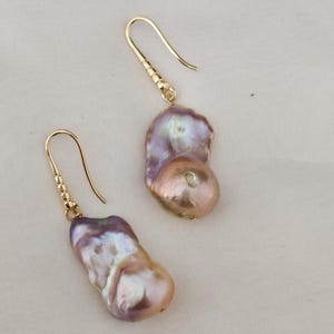 May include: A pair of gold-colored drop earrings featuring large, irregularly shaped, iridescent pearls. The pearls display a blend of colors, including lavender, peach, and cream, with a subtle luster. The earrings have a simple hook design.