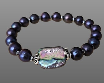 May include: A dark gray pearl bracelet with a large, iridescent, rectangular centerpiece. The bracelet features round, dark gray pearls strung together with silver-toned metal accents. The centerpiece has a unique, textured surface.