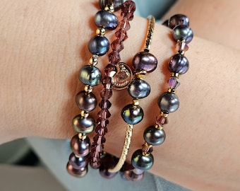 May include: A collection of bracelets featuring dark gray pearls, purple crystals, and gold accents. One bracelet is a gold bangle. The bracelets are stacked on a wrist, showcasing the jewelry's design and color variations.
