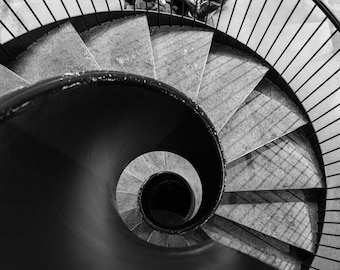 May include: A black and white photo of a spiral staircase with a metal railing. The staircase is made of stone and has a dark, circular opening at the bottom.
