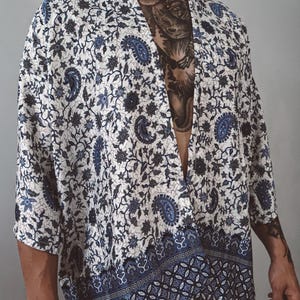 Puede incluir: Una bata tipo kimono y pantalones a juego con estampado de cachemira blanco y azul. La bata tiene un borde ancho con un diseño geométrico. La tela está estampada con motivos de cachemira y florales azules sobre un fondo blanco. El conjunto es ligero y parece estar hecho de una tela suave.
