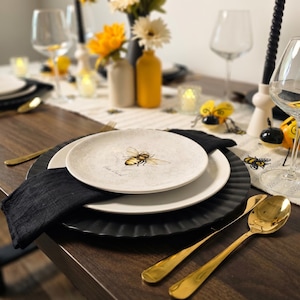 May include: A table setting with a bee theme. Includes white plates with a bee illustration, gold spoons, black napkins, and yellow and white flowers in vases. Wine glasses and candles complete the elegant dining arrangement.