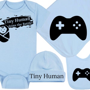 May include: A light blue baby bodysuit, hat, and bib set with a black graphic of a baby and a video game controller. The bodysuit says "Tiny Human Joins the Battle!" and the hat says "Tiny Human".