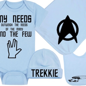 May include: A light blue baby gift set featuring a onesie with the text "MY NEEDS OUTWEIGH THE NEEDS OF THE MANY AND THE FEW" and a Vulcan salute graphic. Includes a matching hat, bib, and blanket with Star Trek-themed designs.