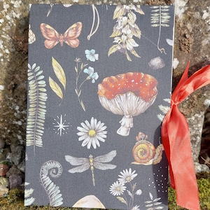 Puede incluir: Un diario gris oscuro con un dise&ntilde;o bot&aacute;nico que presenta una mariposa, una lib&eacute;lula, setas y flores. El diario est&aacute; atado con una cinta naranja brillante. La portada tiene un patr&oacute;n repetido de ilustraciones con temas de la naturaleza.