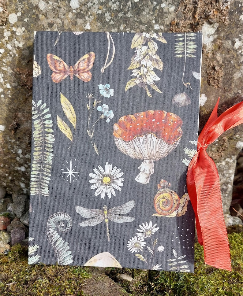Puede incluir: Un diario gris oscuro con un dise&ntilde;o bot&aacute;nico que presenta una mariposa, una lib&eacute;lula, setas y flores. El diario est&aacute; atado con una cinta naranja brillante. La portada tiene un patr&oacute;n repetido de ilustraciones con temas de la naturaleza.