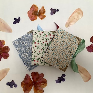 May include: Three small, handmade journals with floral patterned covers. The journals are bound with green ribbon and have gold accents. The journals are arranged in a stack on a white surface with dried flowers scattered around them.