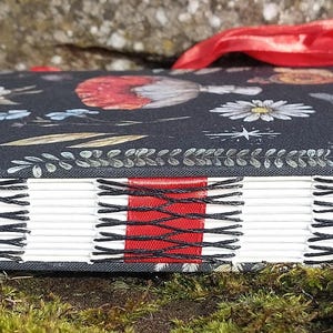 Puede incluir: Un diario hecho a mano con una cubierta de patr&oacute;n floral oscuro con mariposas y flores. Las p&aacute;ginas est&aacute;n encuadernadas con hilo negro y un acento rojo. Una cinta roja est&aacute; unida al lomo.