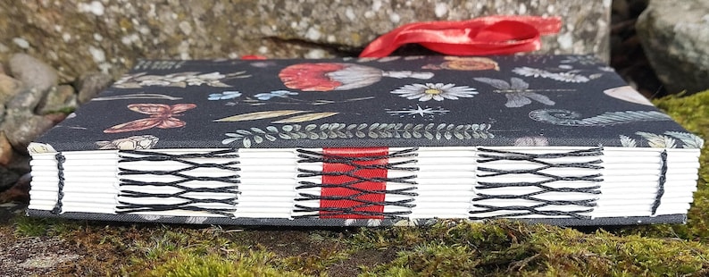 Puede incluir: Un diario hecho a mano con una cubierta de patr&oacute;n floral oscuro con mariposas y flores. Las p&aacute;ginas est&aacute;n encuadernadas con hilo negro y un acento rojo. Una cinta roja est&aacute; unida al lomo.