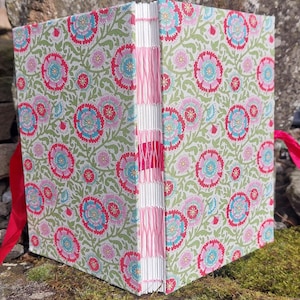 May include: A hand-bound journal with a floral pattern in pink, blue, and green. The cover displays a repeating design of stylized flowers and leaves. The spine is hand-stitched with pink thread, and a red ribbon is attached.