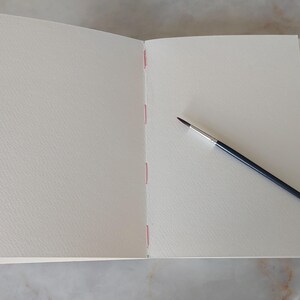 Puede incluir: Un cuaderno de bocetos abierto con p&aacute;ginas en blanco, texturizadas y blanquecinas. La encuadernaci&oacute;n est&aacute; cosida con hilo rosa. Un pincel fino con mango negro y virola plateada descansa sobre la p&aacute;gina derecha. Una cinta roja es visible.