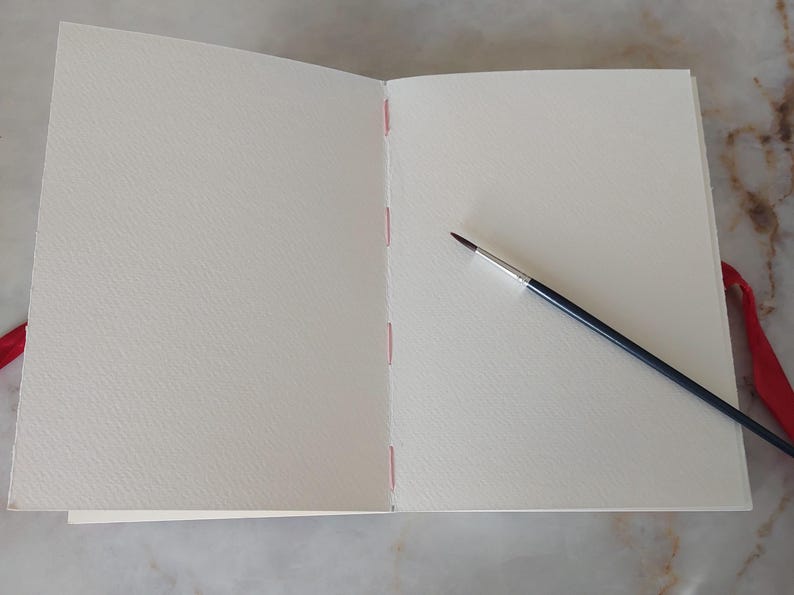 Puede incluir: Un cuaderno de bocetos abierto con p&aacute;ginas en blanco, texturizadas y blanquecinas. La encuadernaci&oacute;n est&aacute; cosida con hilo rosa. Un pincel fino con mango negro y virola plateada descansa sobre la p&aacute;gina derecha. Una cinta roja es visible.