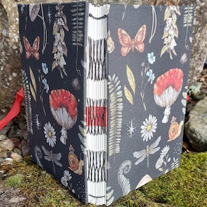Puede incluir: Un diario hecho a mano con una cubierta gris oscuro que presenta un dise&ntilde;o bot&aacute;nico caprichoso. La cubierta est&aacute; adornada con ilustraciones de setas, mariposas, lib&eacute;lulas y flores. El lomo est&aacute; expuesto, mostrando la encuadernaci&oacute;n cosida a mano y una cinta roja.