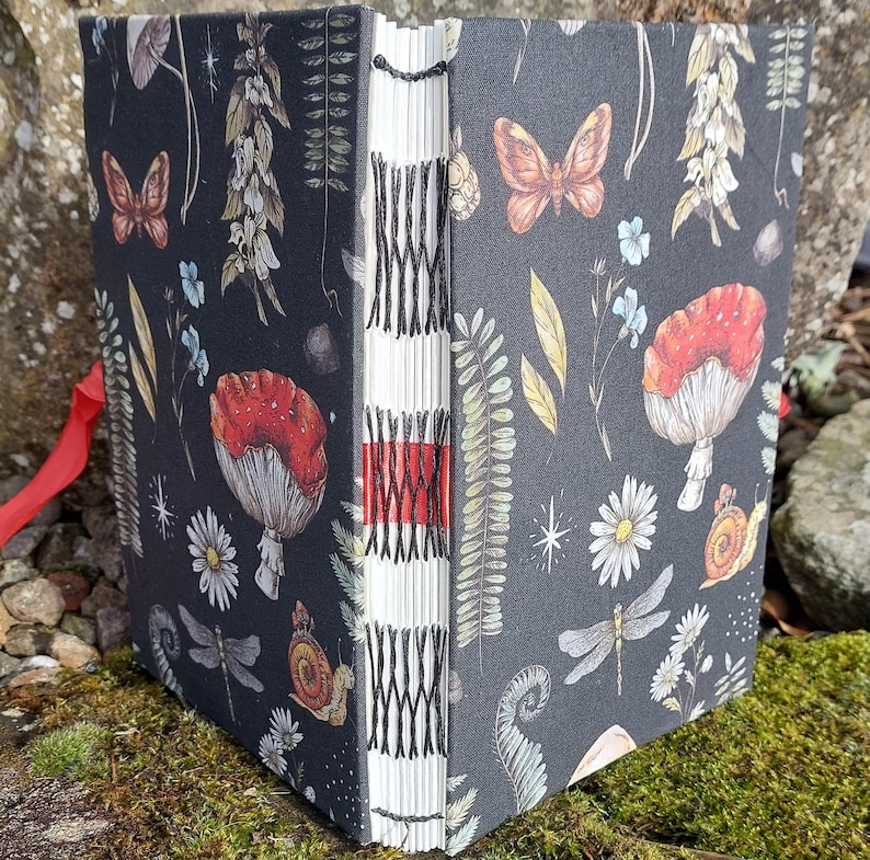Puede incluir: Un diario hecho a mano con una cubierta gris oscuro que presenta un dise&ntilde;o bot&aacute;nico caprichoso. La cubierta est&aacute; adornada con ilustraciones de setas, mariposas, lib&eacute;lulas y flores. El lomo est&aacute; expuesto, mostrando la encuadernaci&oacute;n cosida a mano y una cinta roja.