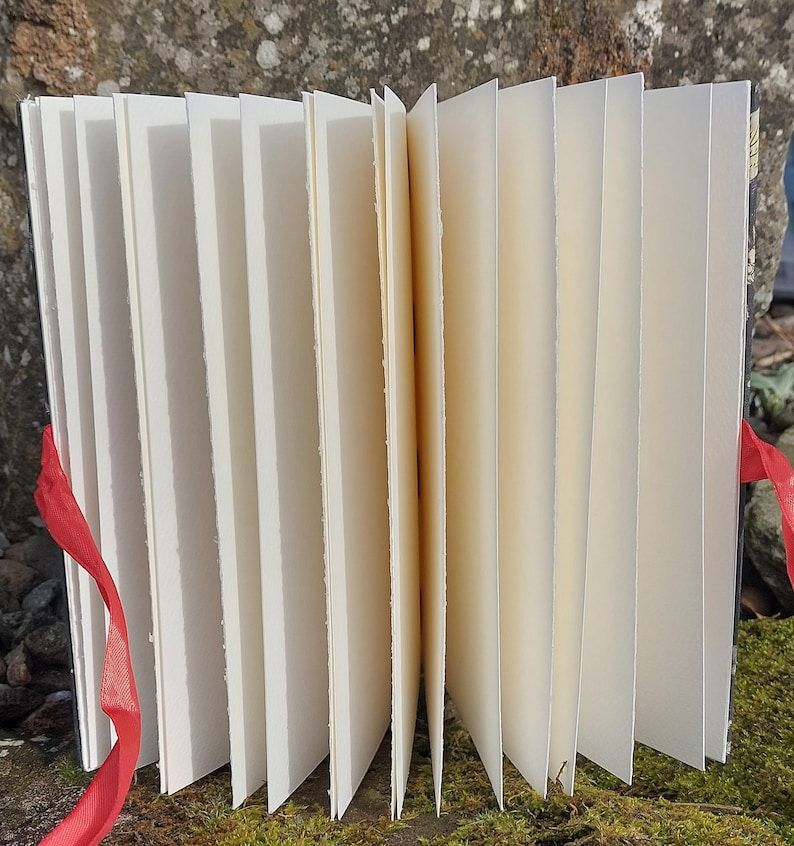 Puede incluir: Un diario hecho a mano, abierto, con p&aacute;ginas color crema. Una cinta roja est&aacute; atada alrededor del lomo. Las p&aacute;ginas est&aacute;n desplegadas, revelando la textura del papel. El diario est&aacute; sobre un fondo natural.