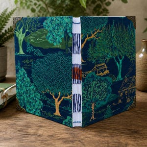 May include: A hardbound book with a dark blue fabric cover featuring a green and teal forest landscape print. The spine is exposed with white stitching and a central orange thread detail. The corners are reinforced with decorative metal accents.