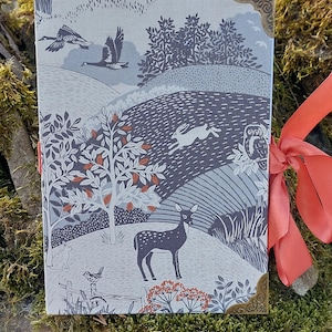 May include: A gray and white journal with a nature-themed design. The cover features a deer, a rabbit, birds, and trees. The journal has a red ribbon tied around it and gold metal corners.