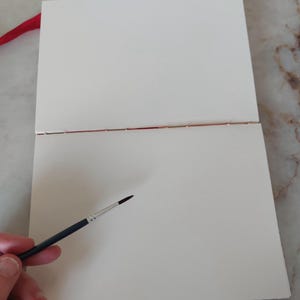 Puede incluir: Un cuaderno de bocetos de acuarela abierto, con p&aacute;ginas blancas texturizadas y una cinta roja. Un pincel con mango negro y punta fina se encuentra cerca de las p&aacute;ginas.