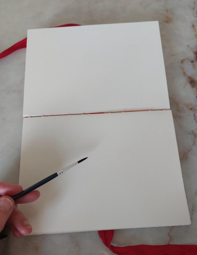 Puede incluir: Un cuaderno de bocetos de acuarela abierto, con p&aacute;ginas blancas texturizadas y una cinta roja. Un pincel con mango negro y punta fina se encuentra cerca de las p&aacute;ginas.