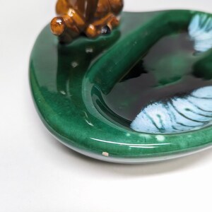 May include: A green ceramic ashtray with a brown ceramic deer figurine sitting on the edge. The ashtray has a white and blue design in the center.