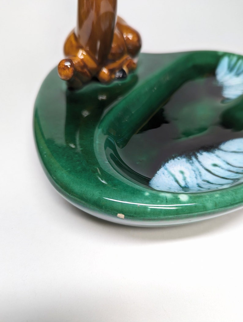 May include: A green ceramic ashtray with a brown ceramic deer figurine sitting on the edge. The ashtray has a white and blue design in the center.