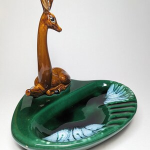 May include: A green ceramic ashtray with a deer figurine on top. The ashtray has a white and green swirl design and a divided section for cigarettes.