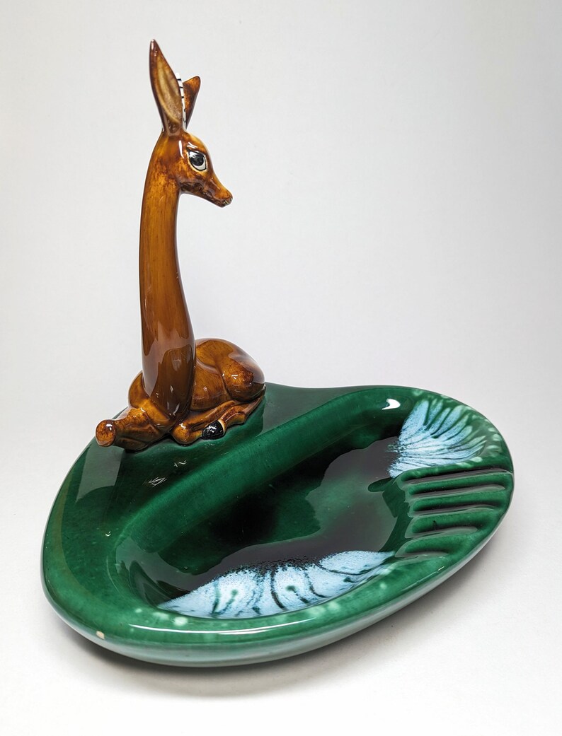 May include: A green ceramic ashtray with a deer figurine on top. The ashtray has a white and green swirl design and a divided section for cigarettes.
