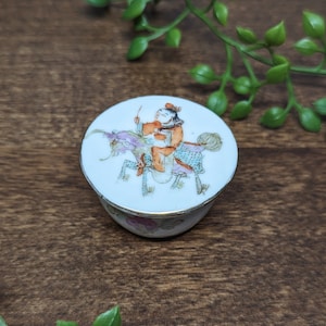 May include: A small, white porcelain trinket box with a round lid. The lid features a colorful hand-painted design of a person riding a mythical creature.
