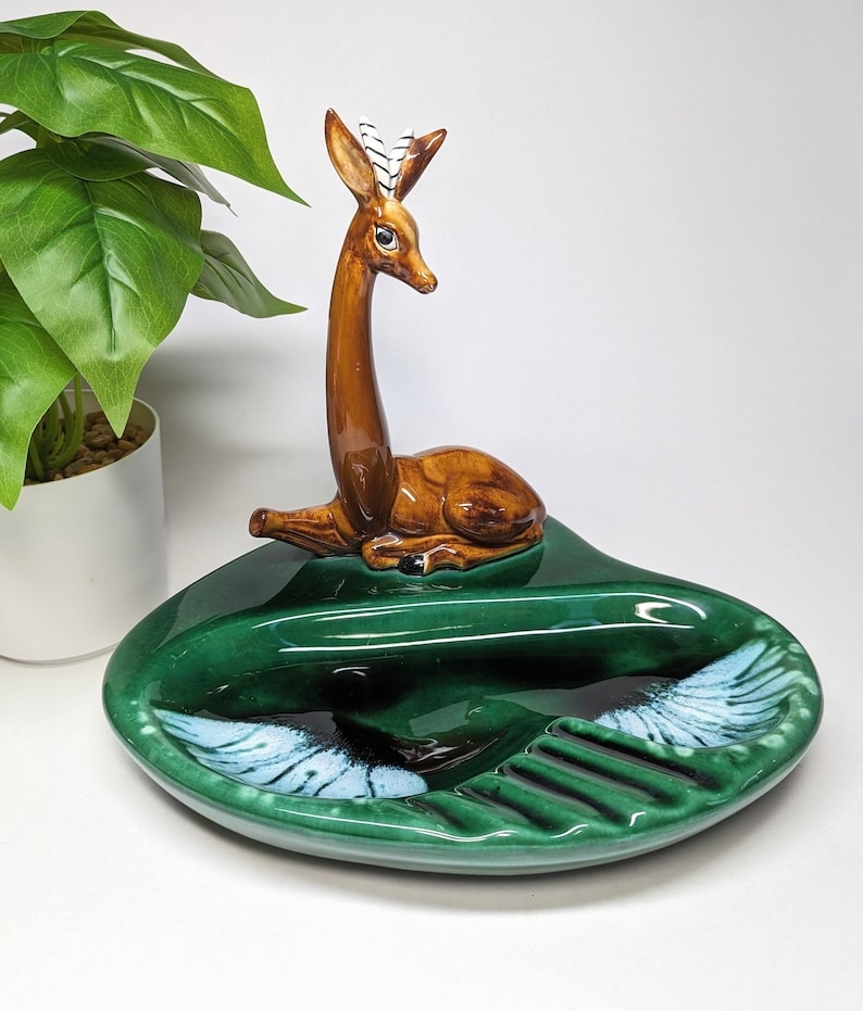 May include: A green ceramic ashtray with a deer figurine on top. The deer is brown with white markings on its antlers. The ashtray has a divided section for cigarettes and a larger section for ashes.